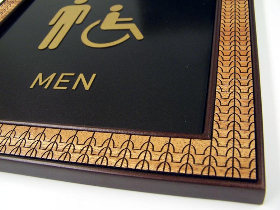 A bathroom sign indicating a men's restroom, featuring gold icons of a standing man and a person in a wheelchair on a black background. This is a closeup of the decorative, patterned frame.