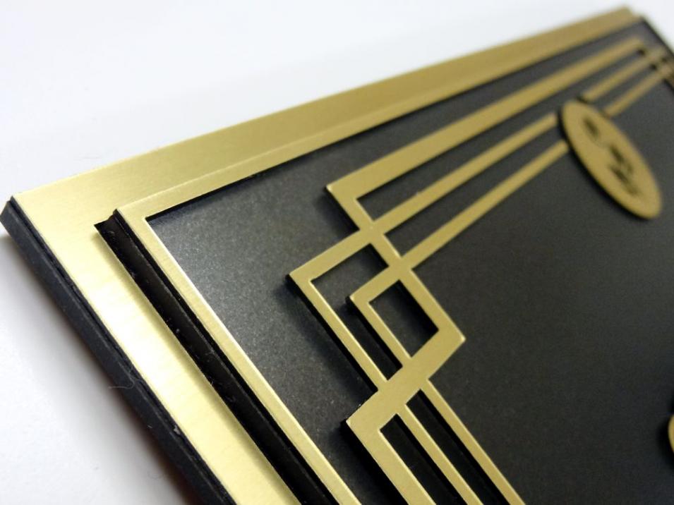 A black sign with gold trim and a gold emblem of leaves at the top center reads "STAFF ONLY" in gold uppercase letters. Below the text, there are tactile braille characters for accessibility. This is a closeup of the border.