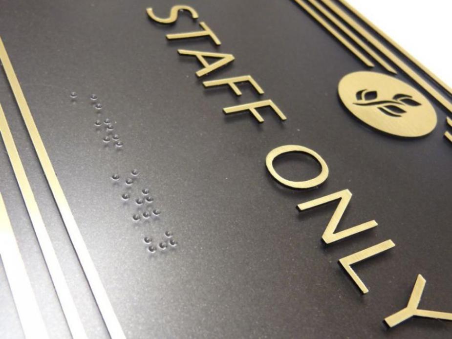 A black sign with gold trim and a gold emblem of leaves at the top center reads "STAFF ONLY" in gold uppercase letters. Below the text, there are tactile braille characters for accessibility. This is a closeup of the raised text and braille.