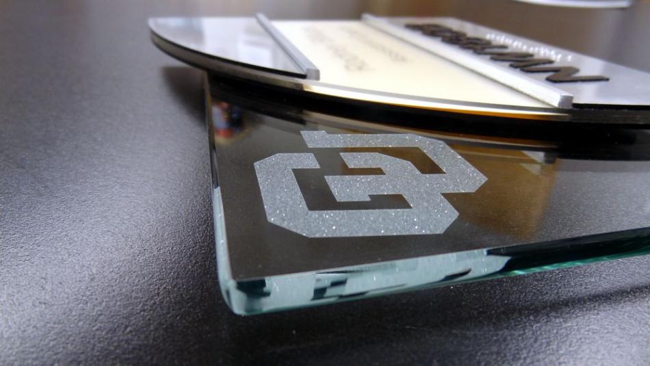 A close-up image of a glass award placed horizontally on a dark surface. The award features an etched logo of an abstract geometric design in the top left corner and layered elements with an engraved text on the top part.