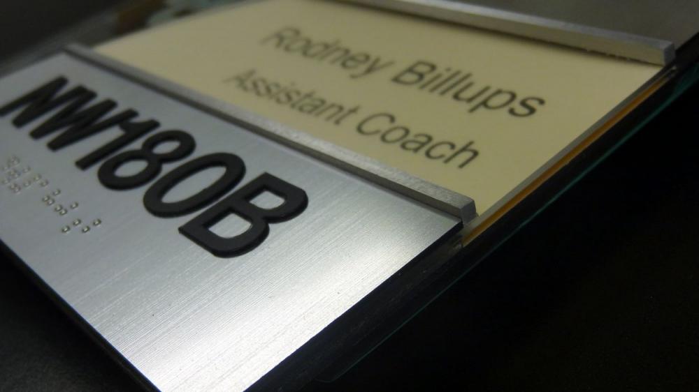 Close-up image of an office nameplate. The nameplate reads "Rodney Billups, Assistant Coach" and includes a raised code "NW180B" beneath the text. The plate appears to be mounted on a wall.