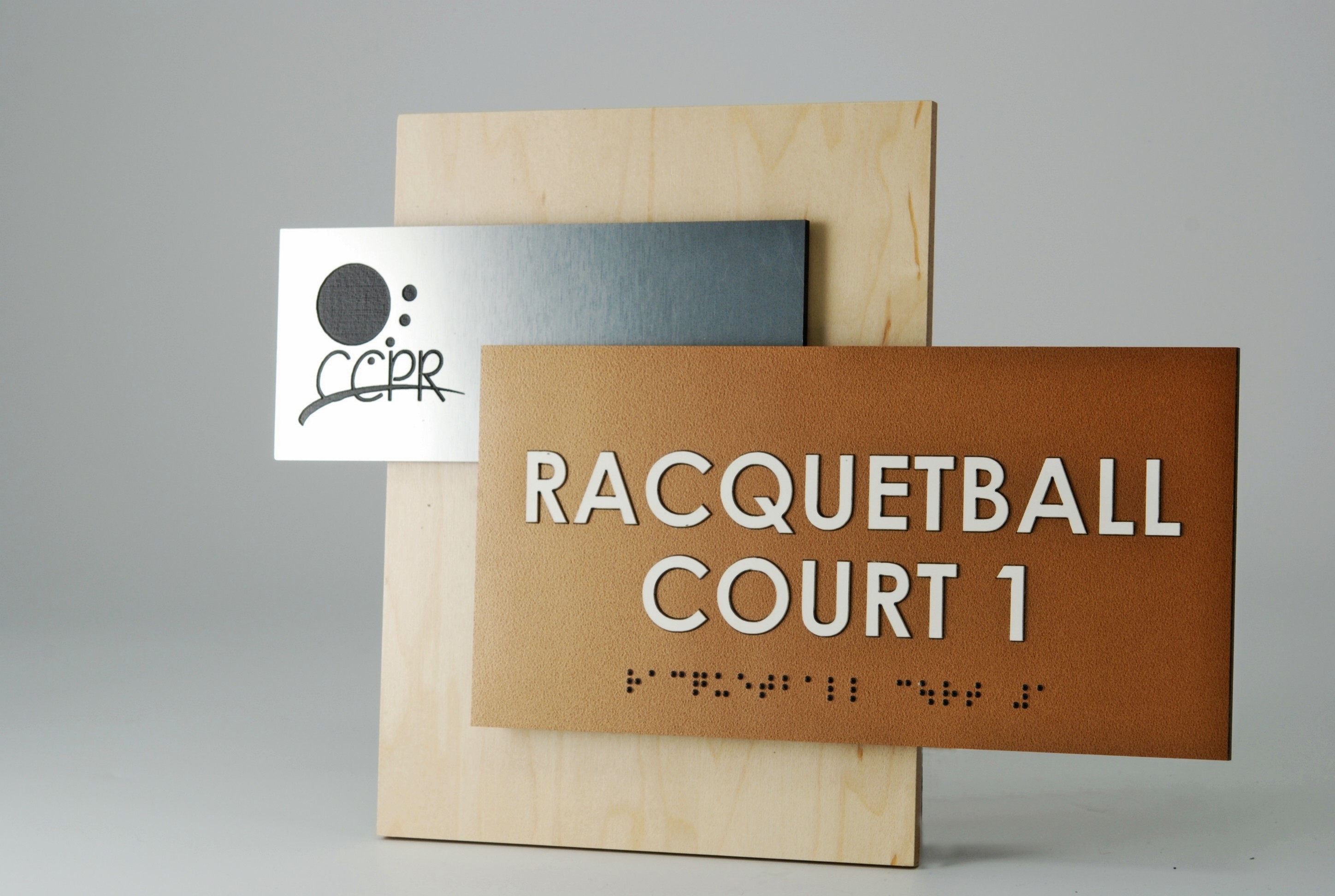 A sign reading "Racquetball Court 1" in large white letters with Braille text below. It is mounted on a wooden board alongside a silver metallic plate with an abstract design and text. The background is plain light gray.
