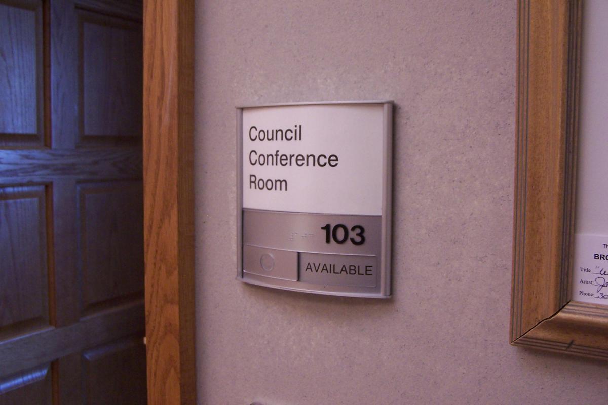 A small rectangular wall sign reads "Council Conference Room" with the number "103" displayed beneath it. The sign is mounted on a light-colored wall, indicating the room's location and purpose within a building.  Raised lettering and braille are present on the metal plate at the bottom of the sign, and a panel has been slid to the left to display text "AVAILABLE".
