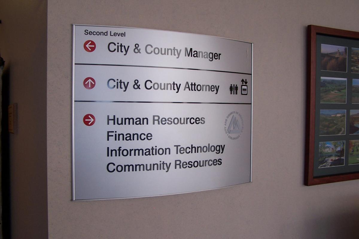 A wall-mounted directory sign inside a building indicates locations for various offices. The second level houses the City & County Manager and City & County Attorney, while the current level has Human Resources, Finance, Information Technology, and Community Resources.