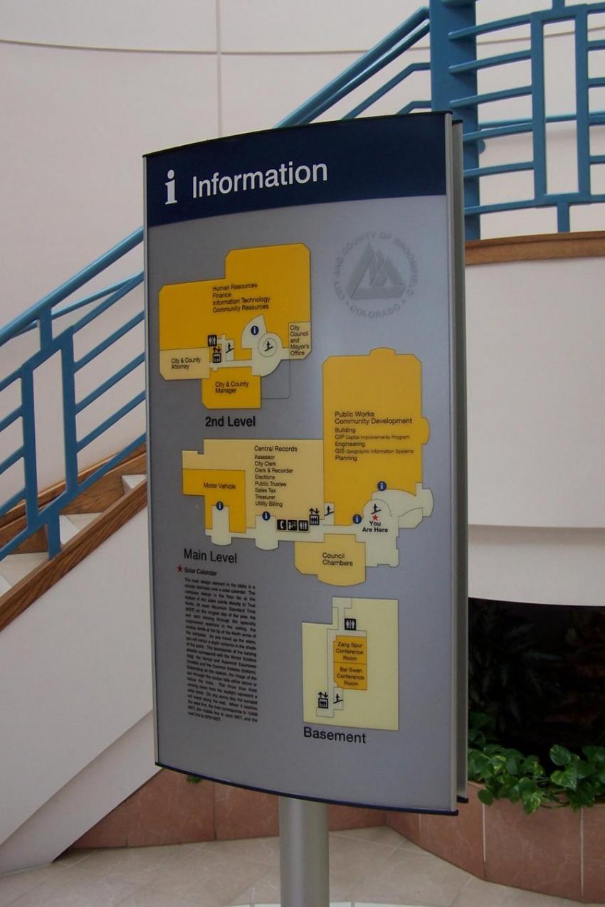 An indoor information sign featuring a map with yellow-highlighted areas labeled "Second Level" and "Basement." It provides details such as directories, restrooms, and points of interest. The sign is located near a staircase with blue railings and plants below.