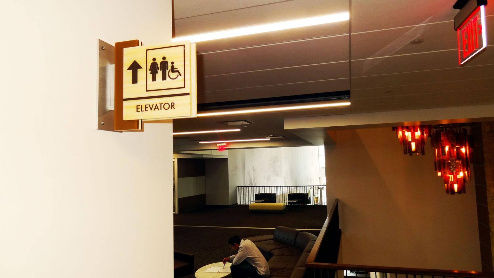 A sign indicating the direction to the elevator is mounted on the wall. The sign includes symbols for an elevator, accessibility for people with disabilities, and directions pointing upwards. Below, a man sits on a chair in a well-lit, modern lobby area.