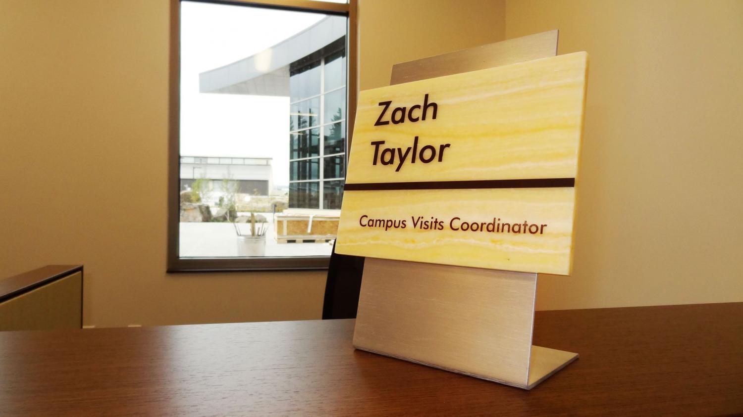 A desk in an office is adorned with a nameplate that reads "Zach Taylor, Campus Visits Coordinator." A large window in the background reveals a modern building exterior with glass features and outdoor landscaping.