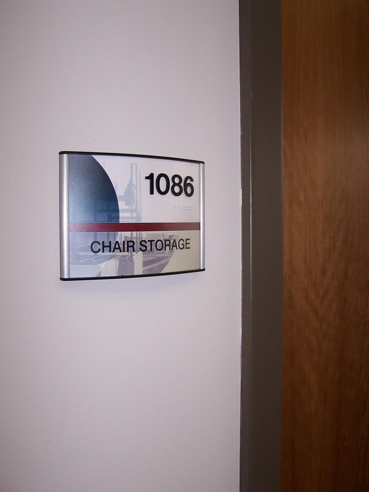 A wall-mounted sign next to a door reads "1086 CHAIR STORAGE." The sign has a silver frame and includes a faintly visible logo in the background. The door beside the sign is wooden with a visible edge. There are subtle shadows around the sign and door frame.