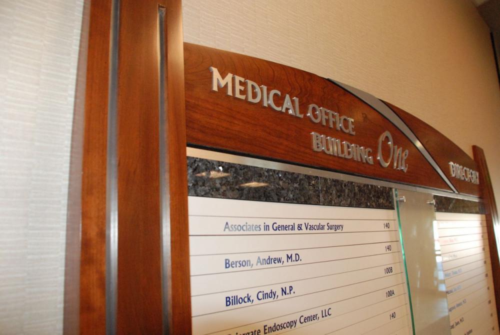 A directory sign inside a building labeled "Medical Office Building One" lists names and room numbers of various doctors. The sign has a wooden frame with a curved top and features a partial glass panel in the center.