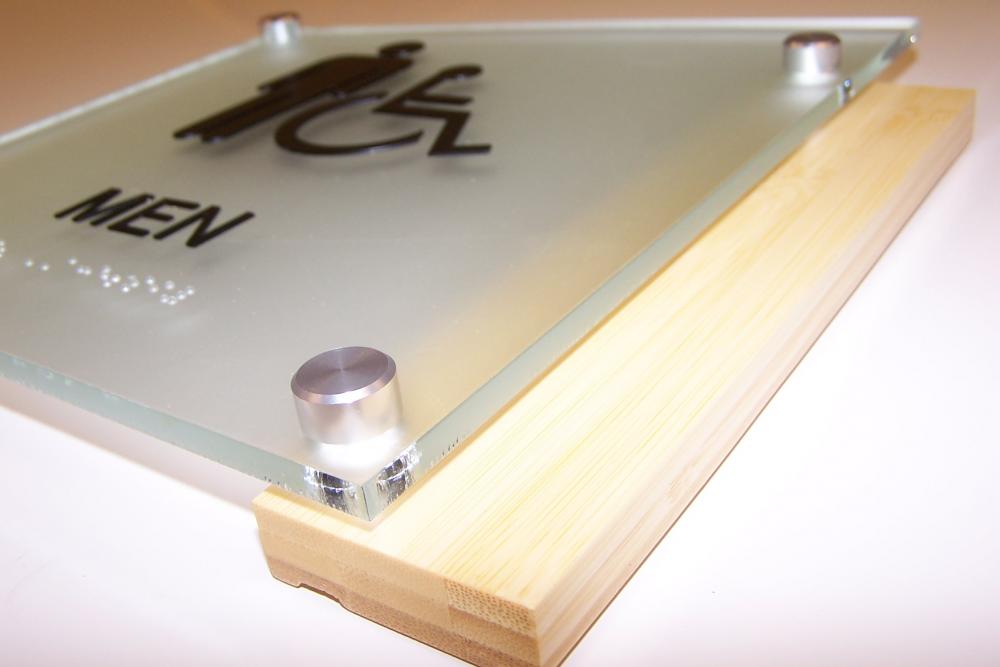 A close-up view of a men's restroom sign featuring a wooden base and a glass top with metal bolts. The sign includes symbols for a man and a wheelchair, indicating accessibility. The word "MEN" is printed below the symbols.