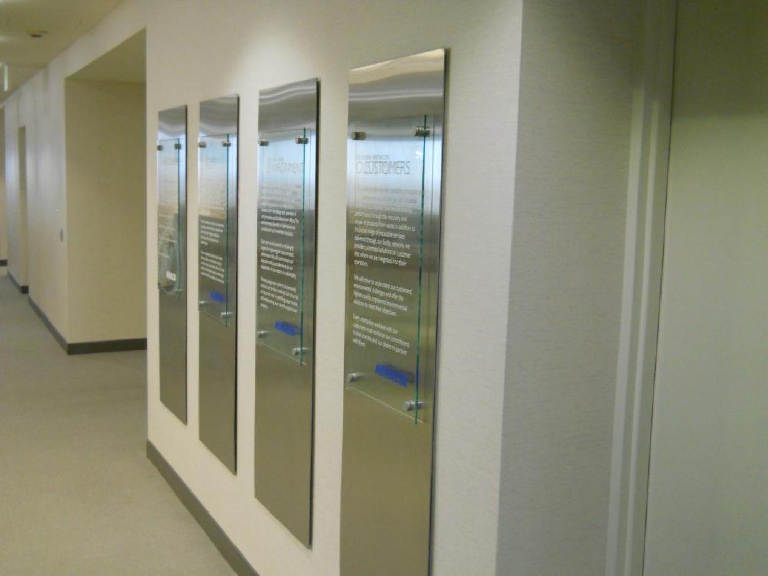 A hallway with four reflective metal plaques mounted on the wall, each etched with text that is partially legible. The plaques are aligned vertically and equally spaced. The wall and floor are light-colored, creating a modern and clean appearance.