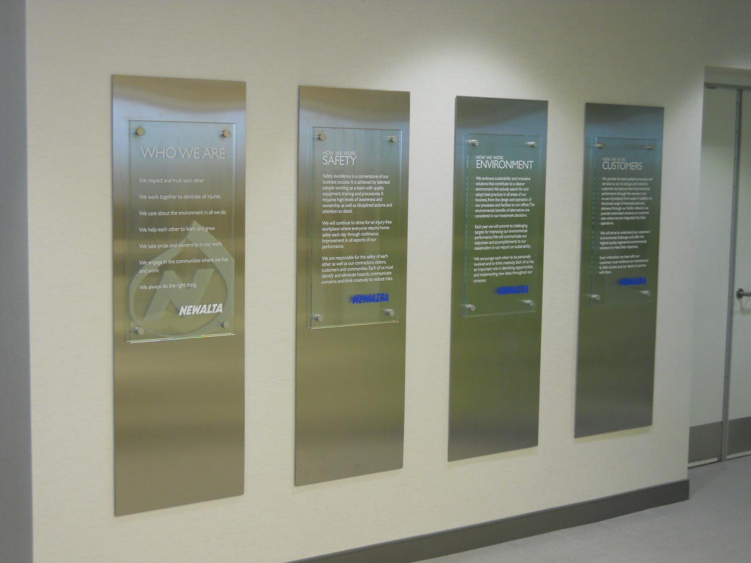 A hallway with four reflective metal plaques mounted on the wall, each etched with text that is partially legible. The plaques are aligned vertically and equally spaced. The wall and floor are light-colored, creating a modern and clean appearance.