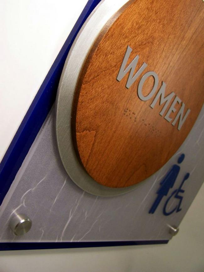 A restroom sign indicating a women's accessible restroom. The sign features a round, wooden plaque with "WOMEN" written in white text, and below it are icons of a female figure and a wheelchair symbol, both in blue. This is a closeup.
