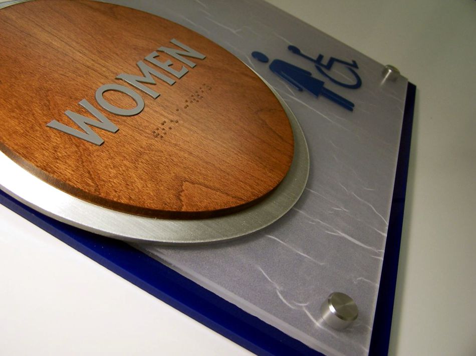 A restroom sign indicating a women's accessible restroom. The sign features a round, wooden plaque with "WOMEN" written in white text, and below it are icons of a female figure and a wheelchair symbol, both in blue. This is a closeup.