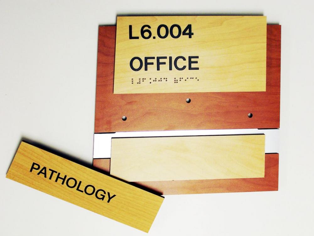 Wooden office signs, one labeled "L6.004 OFFICE" (with braille below it), another labeled "PATHOLOGY." The "OFFICE" sign is mounted on a wooden plaque, while the "PATHOLOGY" sign is placed separately below.