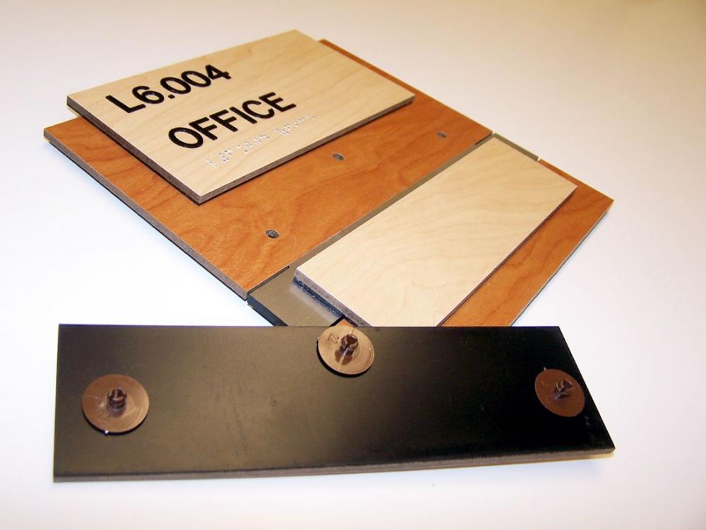A disassembled wooden office sign set, featuring a main board labeled "L6.004 OFFICE" along with tactile Braille and a mounting bracket with three fasteners. The components are displayed on a plain white surface.