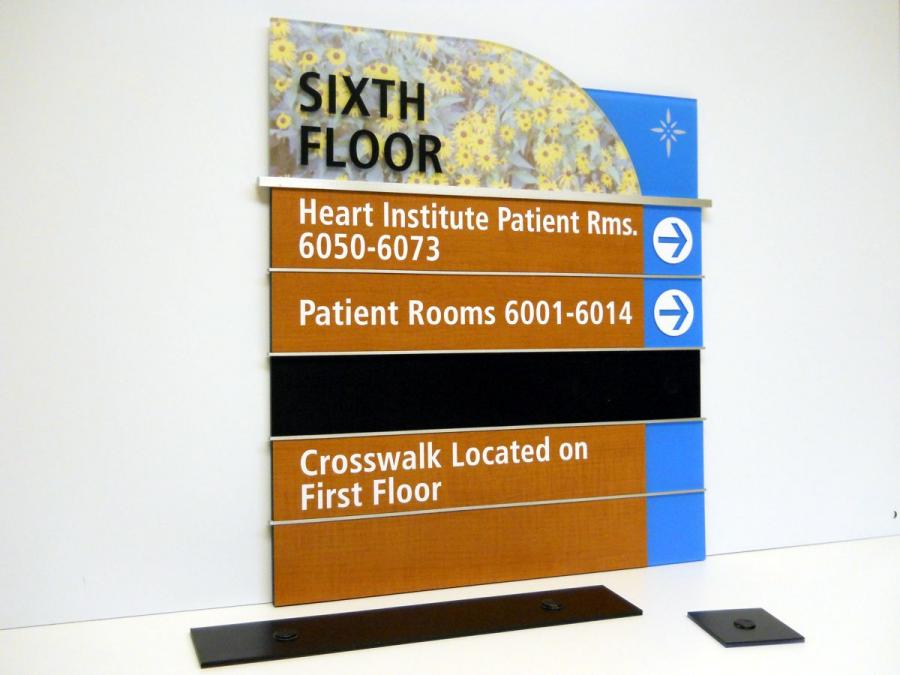 A hospital directory sign for the sixth floor showing directions. It lists: "Heart Institute Patient Rms. 6050-6073," "Patient Rooms 6001-6014," both with arrows pointing to the right, and "Crosswalk Located on First Floor." The sign has floral and blue decorative accents.