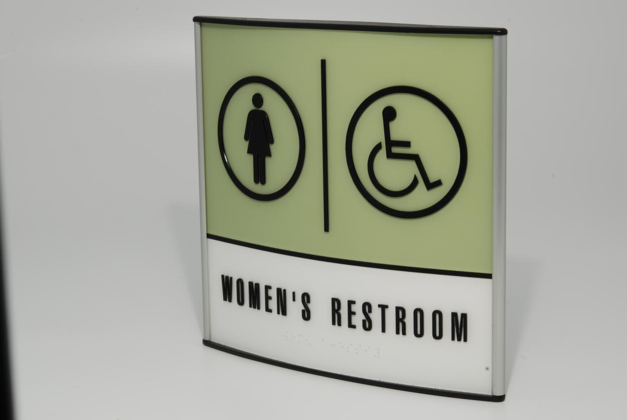 A rectangular sign indicating a women's restroom, featuring a female figure icon on the left and a wheelchair accessibility icon on the right. The background is light green, with the text "WOMEN'S RESTROOM" in black letters at the bottom.