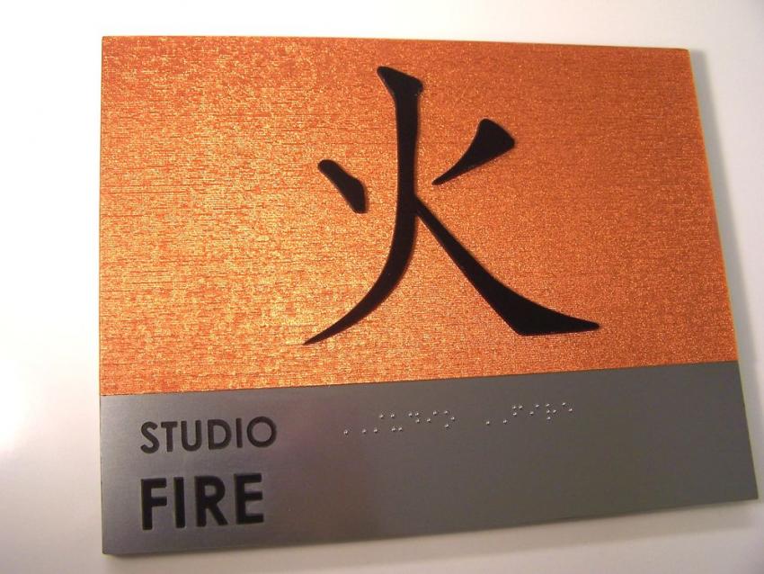 A sign with a metallic orange background features a large black kanji character for "fire." Below, on a silver and gray section, the text reads "STUDIO FIRE" in black capital letters. There is also a section with Braille text below the written text.