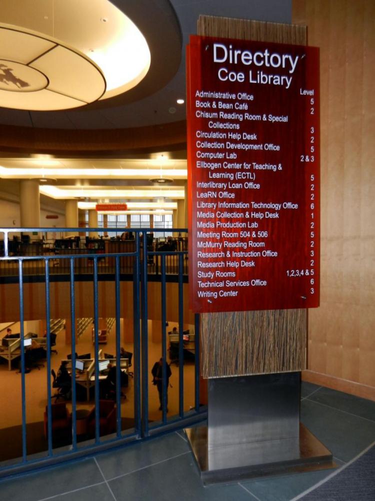 A vertical red directory sign with white text stands on a metal base in a spacious, modern building with multiple floors. The sign lists the locations of various offices and facilities within the Coe Library and features a wooden accent at the bottom.
