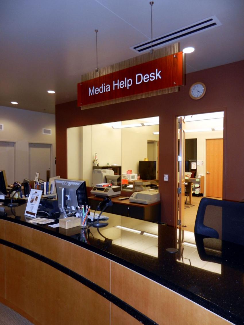 A media help desk with a counter featuring computers, office supplies, and a sign overhead reading "Media Help Desk." Behind the counter is an office area with a clock on the wall and various office equipment, visible through a large window cut-out.
