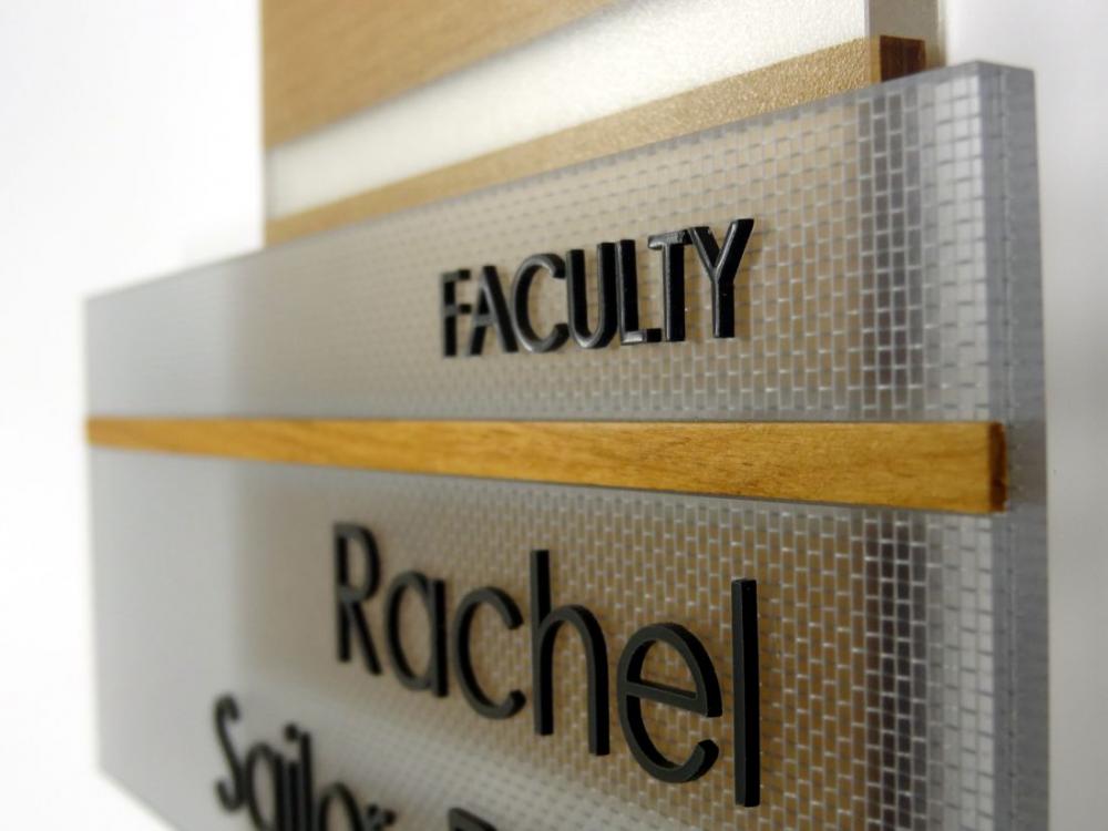 A textured sign with the word "FACULTY" in bold black letters at the top. Below it, separated by a wooden strip, is the name "Rachel" followed by partially visible text "Sailor". The background consists of frosted and clear panels.