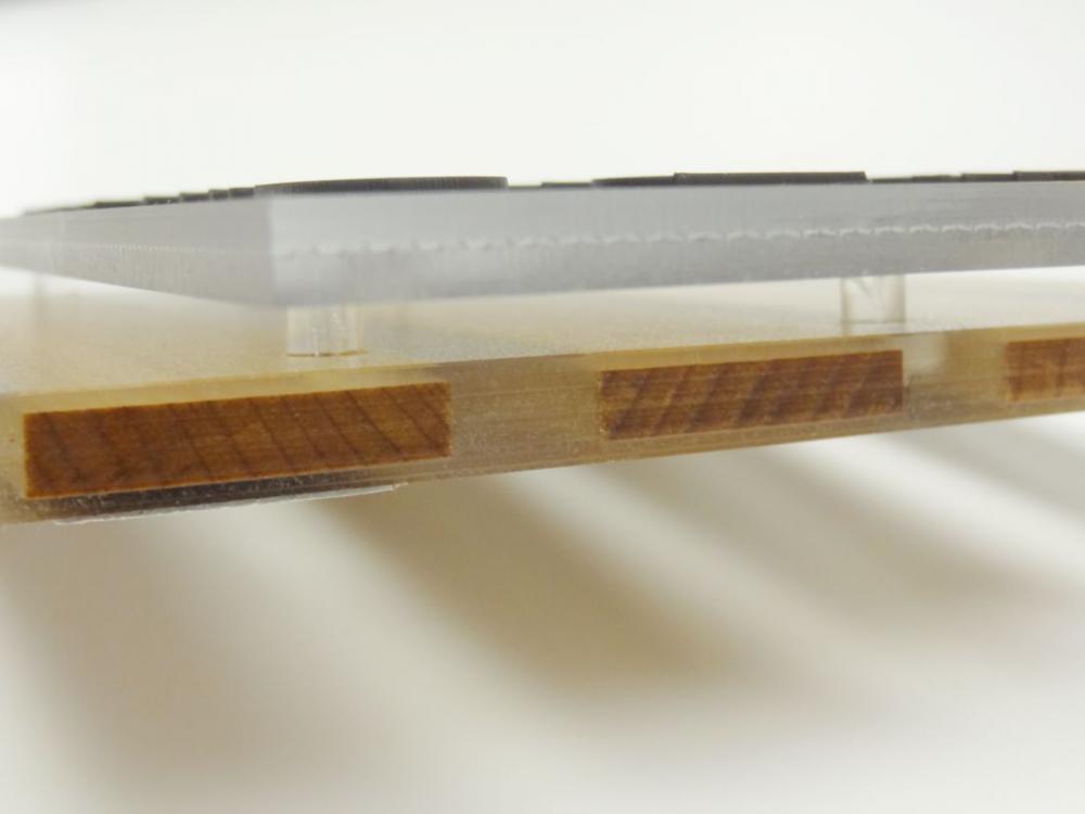 Close-up of a layered wall sign, where a clear plastic layer is placed on top of a wooden layer. The plastic layer is elevated above the wooden layer by small clear supports.