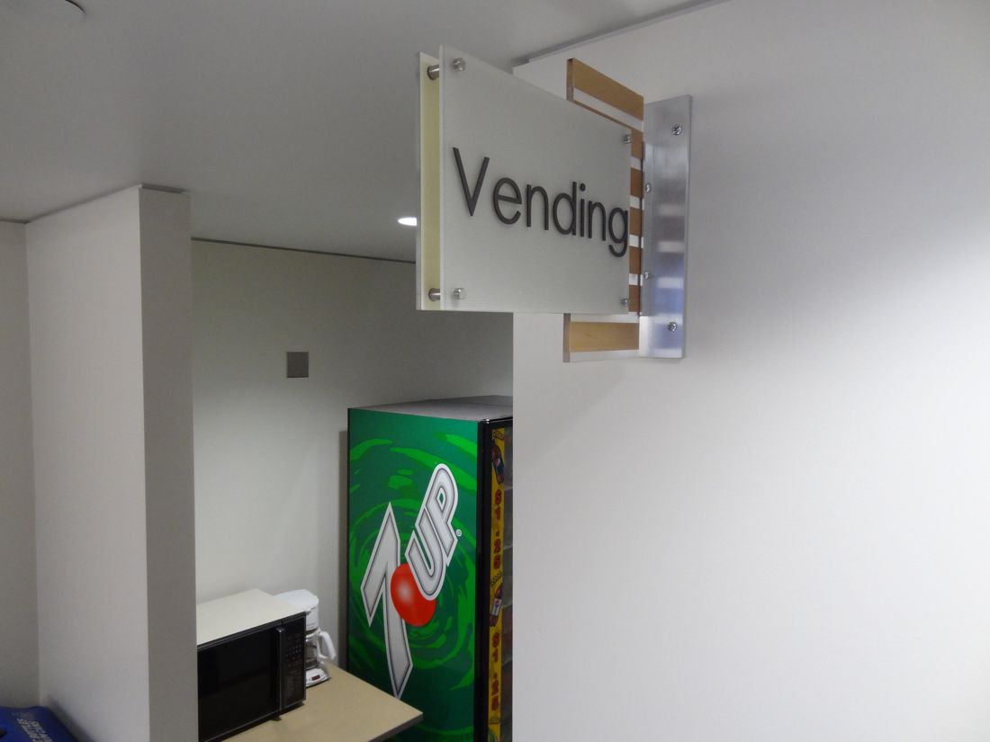 A vending area with a sign that reads "Vending" mounted on the wall. Below the sign, there is a green 7UP vending machine with a red button design. A microwave oven and a countertop are partially visible next to the vending machine.