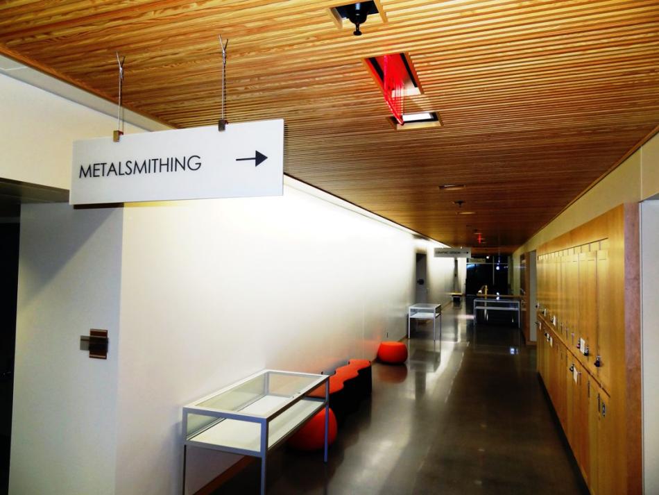 A hallway with a wooden ceiling and lockers on the right side. A sign hanging from the ceiling indicates the direction to a "Metalsmithing" room with an arrow pointing forward. There are glass display cases and orange stools along the left wall.