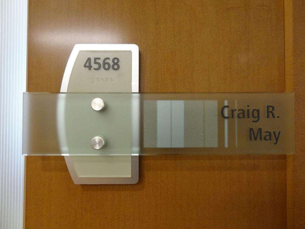 A modern office door sign on a wooden panel features the name "Craig R. May" and the office number "4568." The sign combines metallic elements with frosted and transparent glass panels, including Braille.