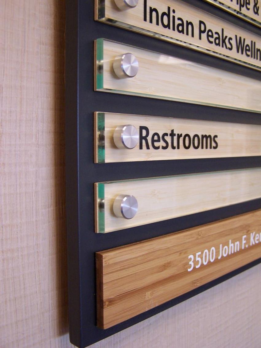 Close-up of a directory sign with sections labeled as "Indian Peaks Welln..." and "Restrooms" among others. The sign has multiple wooden panels with metal fasteners and clear separators, and the address "3500 John F. Ken..." is partially visible at the bottom.