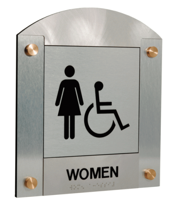 A silver restroom sign with braille text at the bottom. The sign features a symbol of a woman and a wheelchair user, indicating that the restroom is accessible for women and individuals with disabilities.