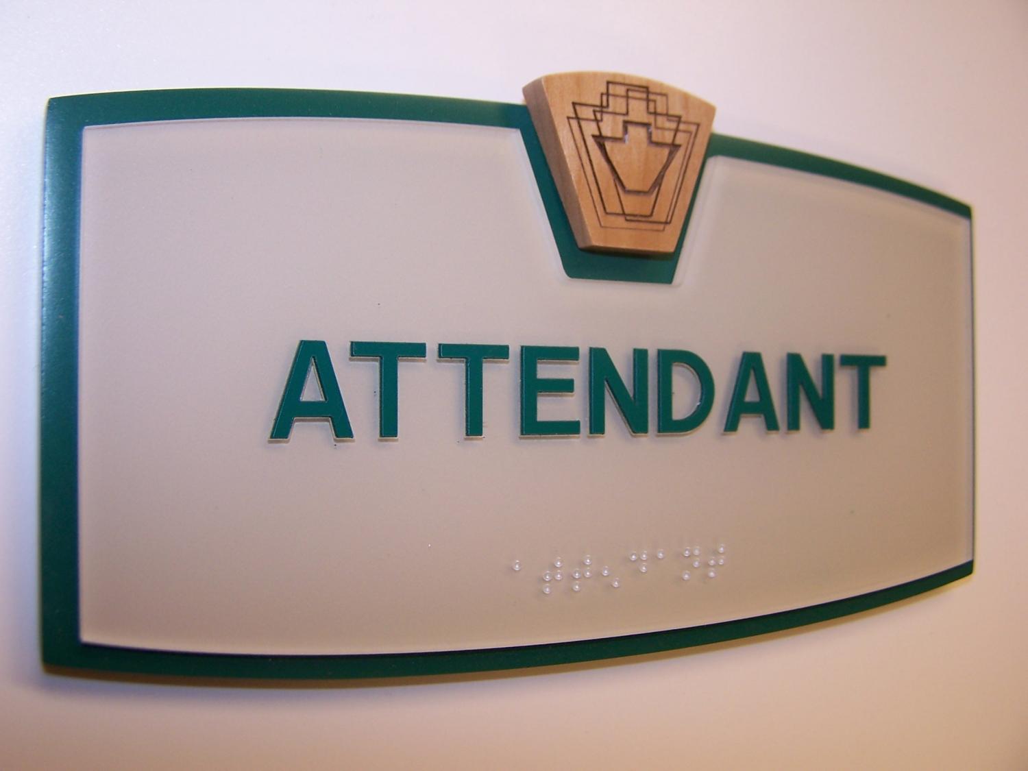 A frosted glass sign with green borders and a wooden emblem at the top, displaying the word "ATTENDANT" in green uppercase letters. Below the text, there is braille writing. The sign is mounted on a white wall.
