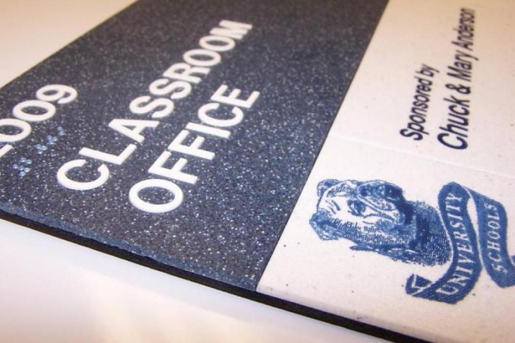 Close-up of a sign indicating a "Classroom Office" for room 2009, sponsored by Chuck and Mary Anderson. The sign features a blue and white color scheme and includes an icon of a bulldog wearing a University Schools uniform.