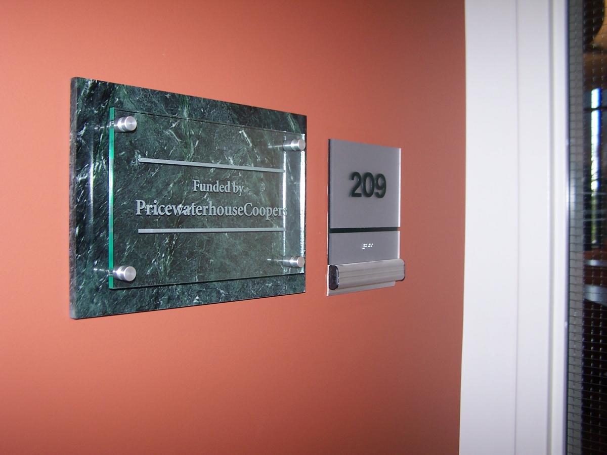 A maroon-colored wall with a sign that reads "Funded by PricewaterhouseCoopers" mounted on a green marble-looking background with clear panels held by metallic supports. Next to it is a room number plaque displaying "209" with braille underneath.