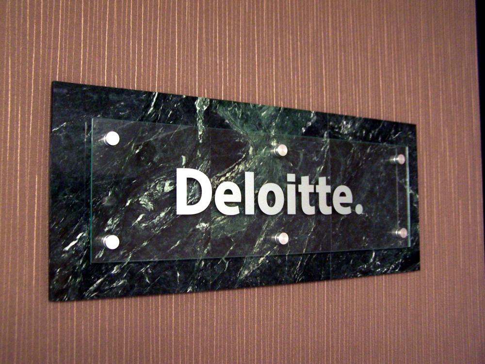 A rectangular sign with the text "Deloitte" in white letters is mounted on a dark green, marble-like background with four silver fasteners at the corners. The sign is affixed to a vertically striped brown wall.