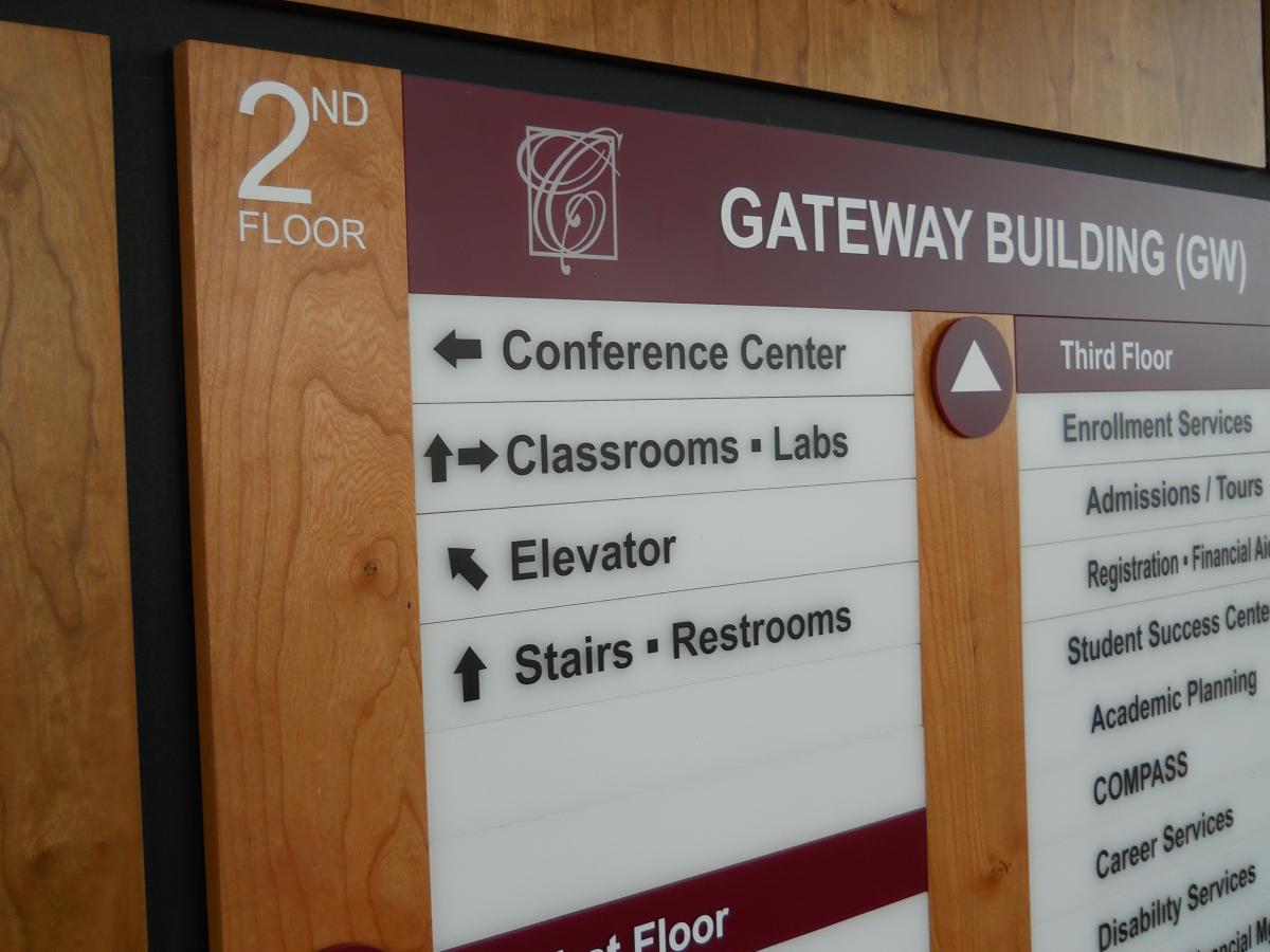 A wood paneled wall featuring a  directory board. The directory lists various departments and their locations across four floors, including classrooms, labs, and student services.  This is a closeup of the signage.
