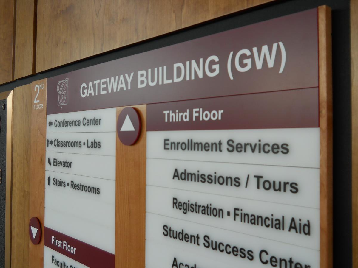 A wood paneled wall featuring a  directory board. The directory lists various departments and their locations across four floors, including classrooms, labs, and student services.  This is a closeup of the signage.