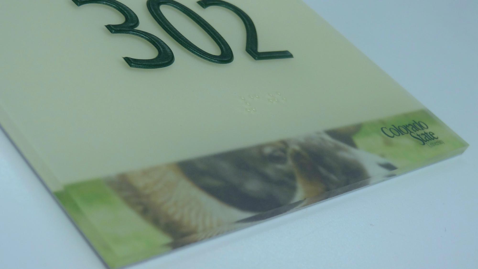 A room number sign displays the number "302" in raised green font. The lower part of the sign features a partial image of a ram and includes the "Colorado State" logo.  This is a closeup that also shows the braille text below the numbers.
