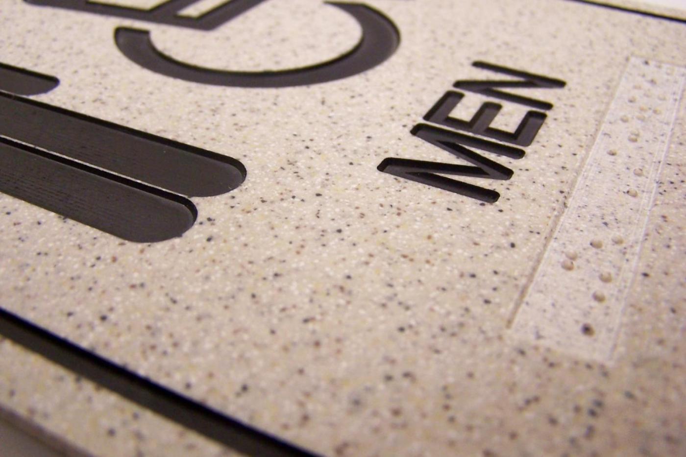 A close-up view of a textured restroom sign for men. The sign has raised black text on a pale, speckled background, and the word "MEN" is clearly visible. Braille dots are also present below the text for accessibility.