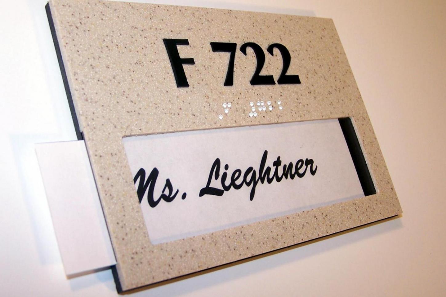 A door sign with large text "F 722" at the top, and text in Braille beneath it. Below, there's a clear window displaying a piece of paper with the handwritten name "Ms. Lieghtner." The sign is mounted on a light-colored wall.