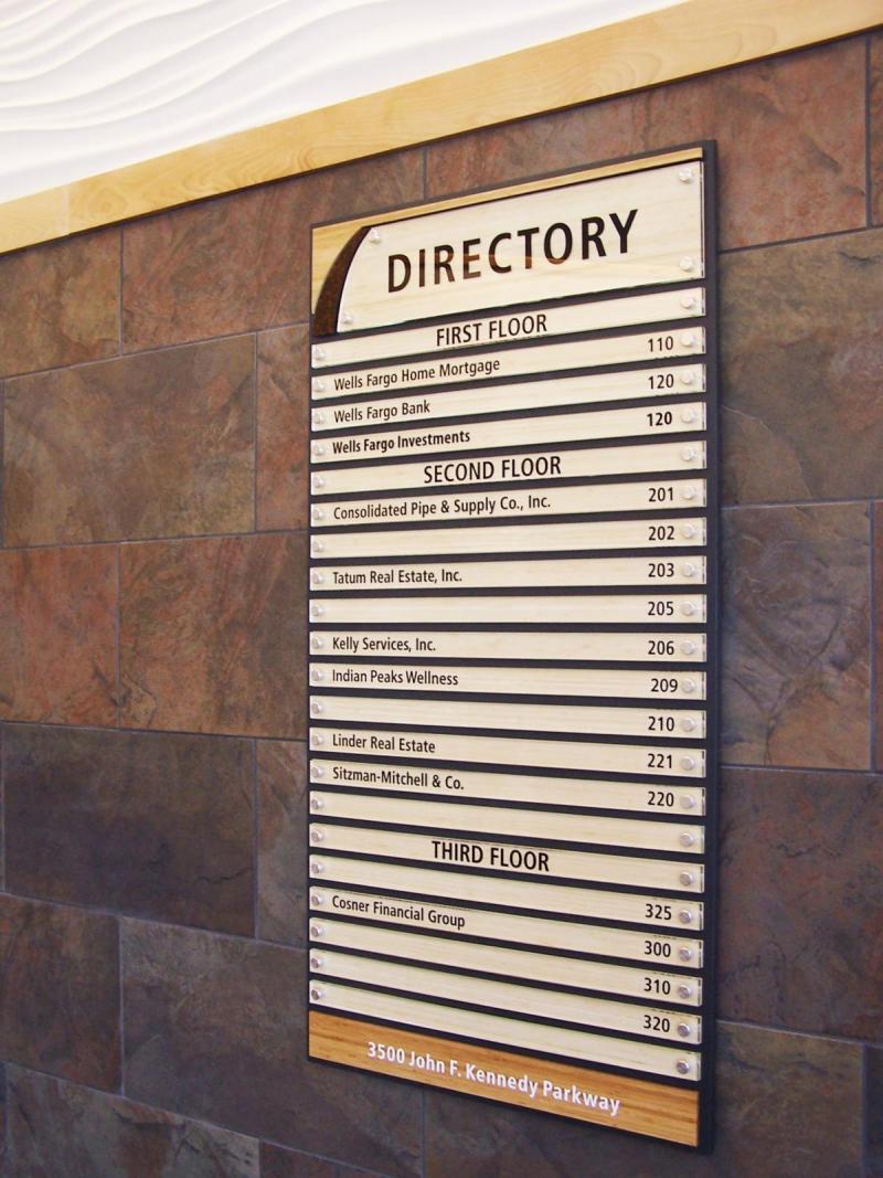A building directory showing listings for three floors. The first floor includes Wells Fargo Home Mortgage (110), Wells Fargo Bank (120), Wells Fargo Investments (130). The second floor lists various companies, and the third floor lists Cosner Financial Group (325) and Cosner Financial (330). The directory is mounted on a wall with tile and wood accents. The bottom includes the address: 3500 John F. Kennedy Parkway.