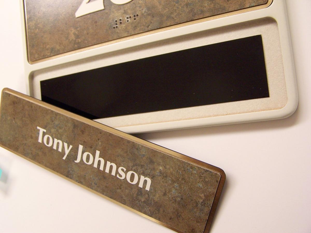 A nameplate reading "Tony Johnson" lies diagonally across an empty nameplate slot mounted on a wall. The surface of the nameplate has a marble-like texture with white text, and the slot is outlined in white.