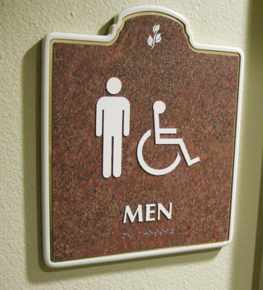 A restroom sign on a wall indicating a men's restroom. The sign features icons of a male figure and a person in a wheelchair, along with the word "MEN" beneath them. The sign has a brown background with white text and border, and raised braille text