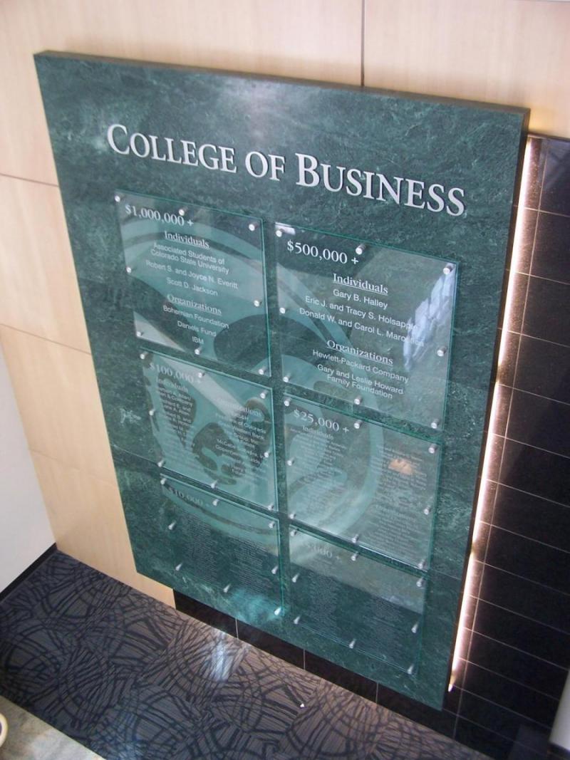 An interior display at a university's College of Business features a tall, green marble-like plaque with glass panels listing names. The backdrop includes wooden paneling.