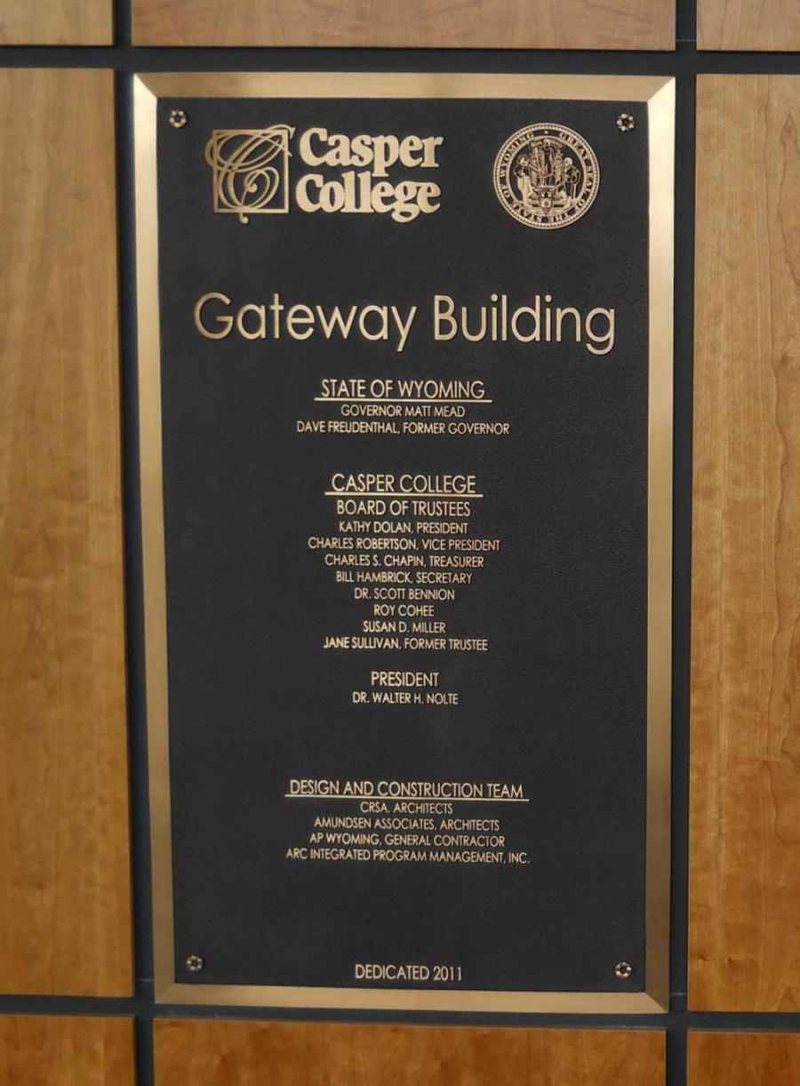 A bronze plaque mounted on a wooden surface, commemorating the "Gateway Building" at Casper College. It lists state officials, the Board of Trustees, the president, and the design and construction team. It is dedicated in 2011.