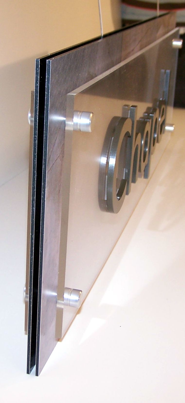 A sign mounted on a wall reads "Chapel" in bold, raised lettering. The background of the sign has a subtle, earthy-toned pattern with a rectangular shape and secured at the corners with visible screws.