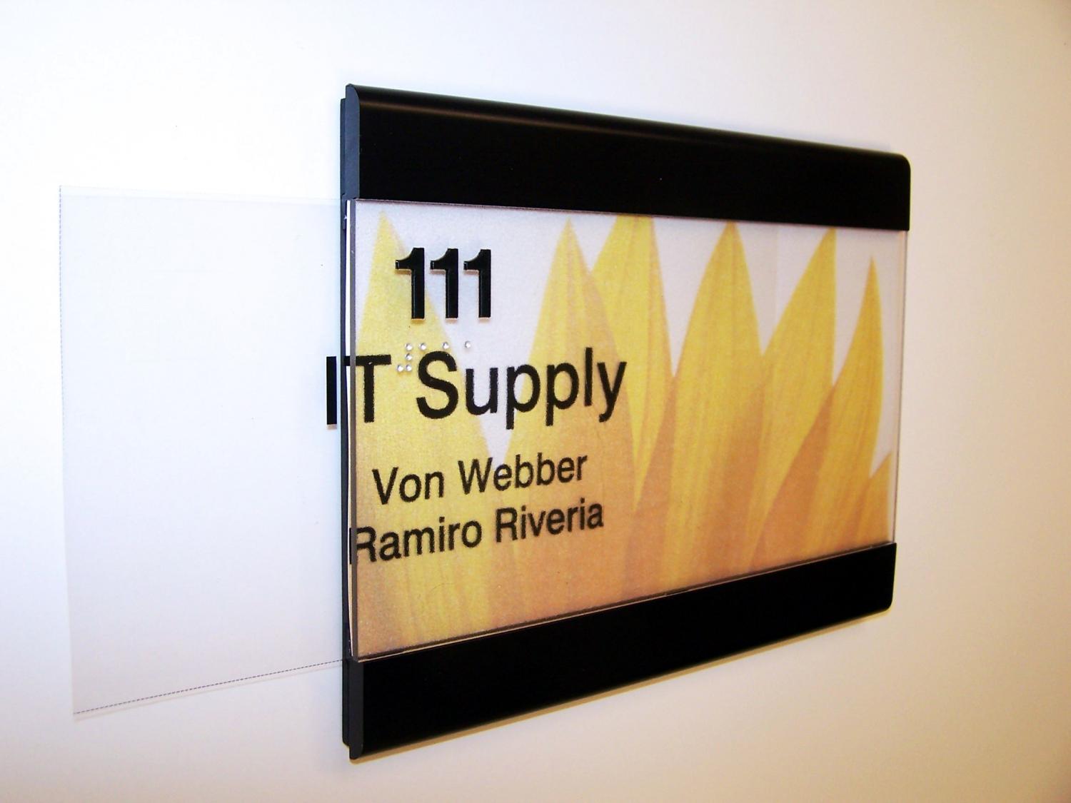 A sign on a wall displays "111 IT Supply" in black text against a background of yellow petals. Below, it lists the names "Von Webber" and "Ramiro Riveria." The sign has black borders at the top and bottom, with a transparent side panel extending out to show that it is interchangeable.