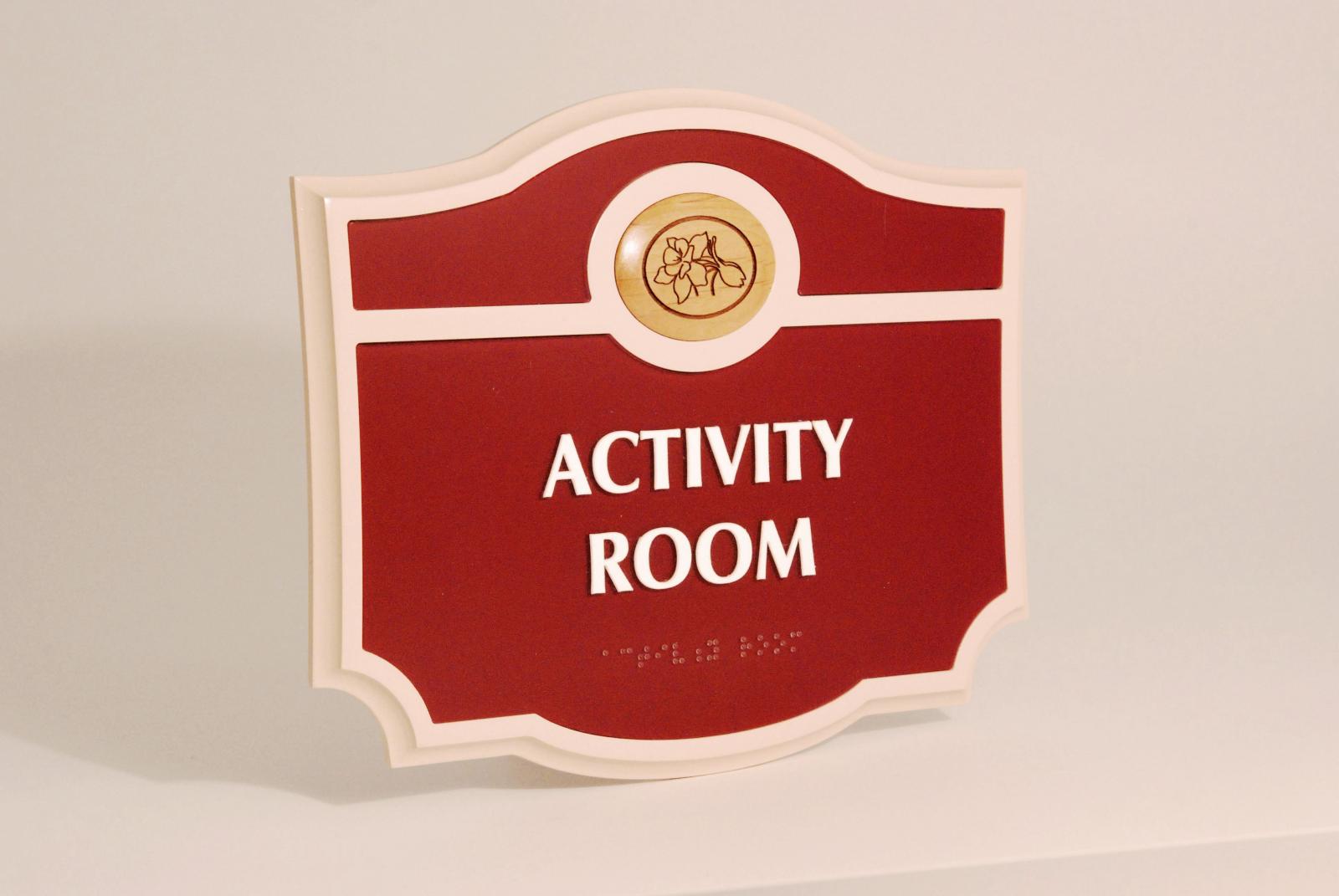 A red and white sign with an ornate shape, displaying the words "ACTIVITY ROOM" in bold white letters. The top part of the sign features a small circular emblem with an illustration of a flower inside it.