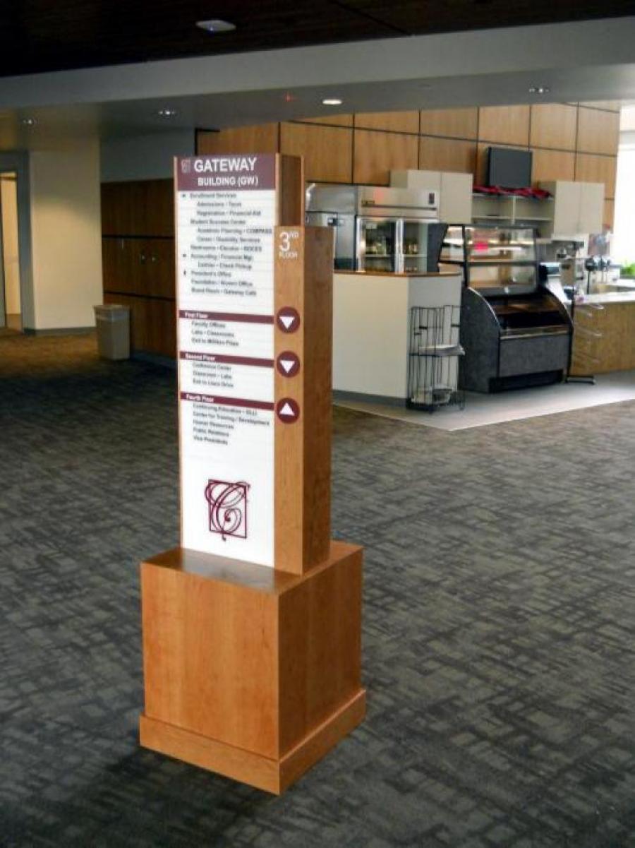 A directory sign in a building hallway indicating locations for various departments and offices under "Gateway Building (GW)." The sign has arrows pointing to different floors, a graphic at the bottom, and double doors in the background.